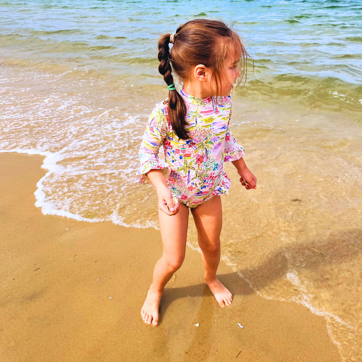 Child in a floral swimsuit standing on a beach with ocean and boats in the background