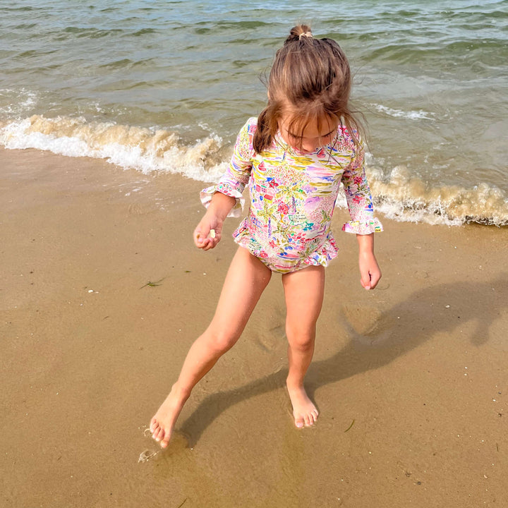 Child in a floral swimsuit walking on a sandy beach with ocean waves.